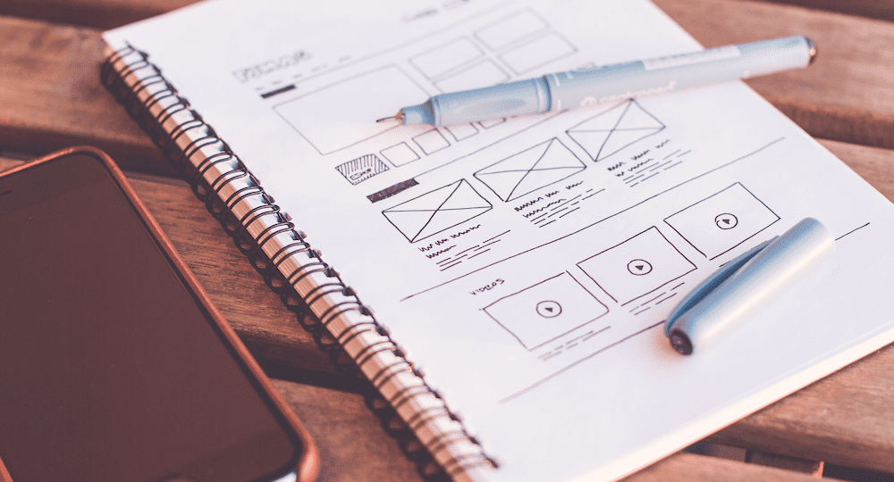 laptop and notebook sitting on a wooden table with a wireframe drawn in the notebook with a pen