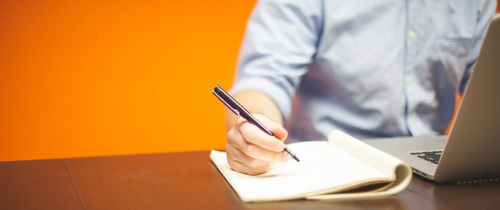 Man in front of orange wall with a laptop writing in a notebook